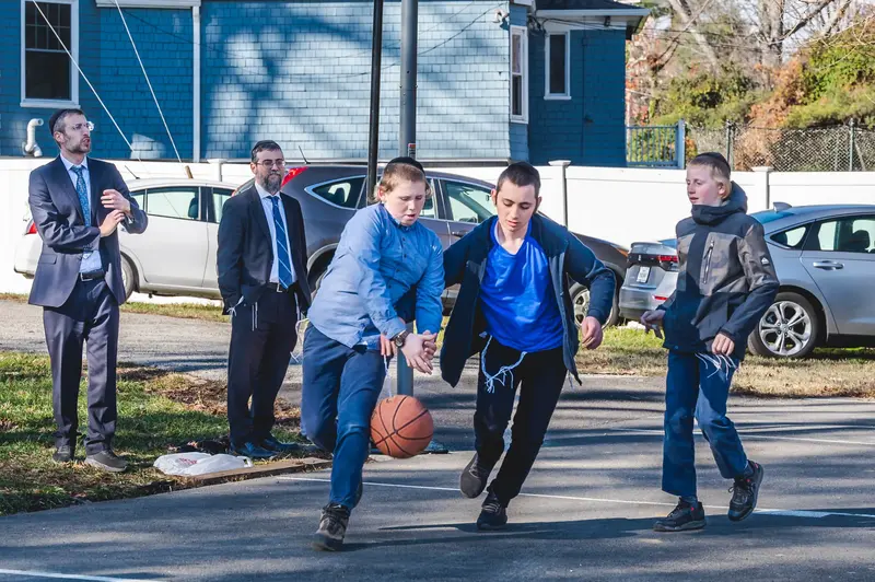 Boys playing basketball on the court at recess while two men in suits watch from the sideline