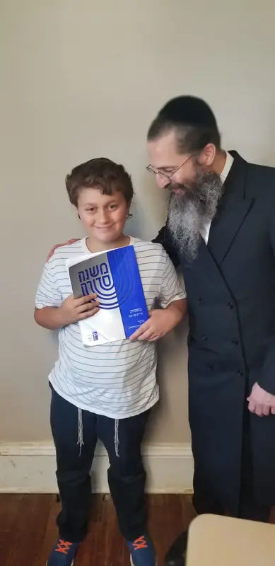 A smiling boy holding a Mishneh Torah volume beside a Rebbi