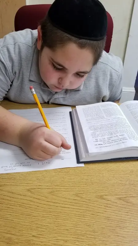A boy writing answers on a worksheet next to an open Hebrew sefer