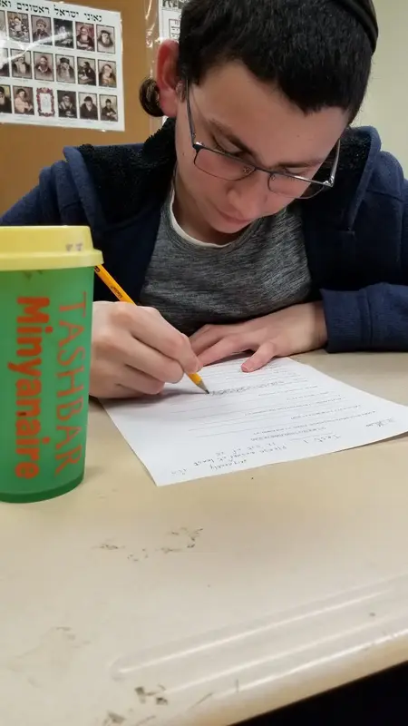 A student working on a worksheet beside a green Tashbar Minyanaire cup