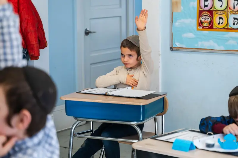 A boy raising his hand at his desk in a classroom with an Alef-Beis poster on the wall