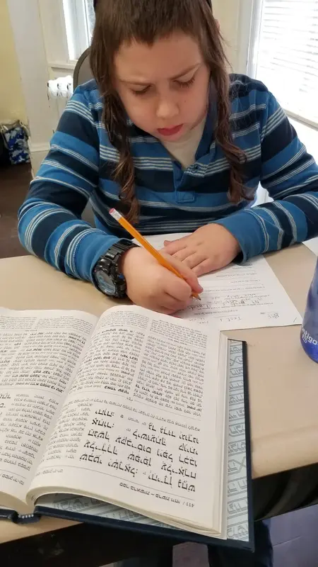 A boy writing on a worksheet beside an open Hebrew text
